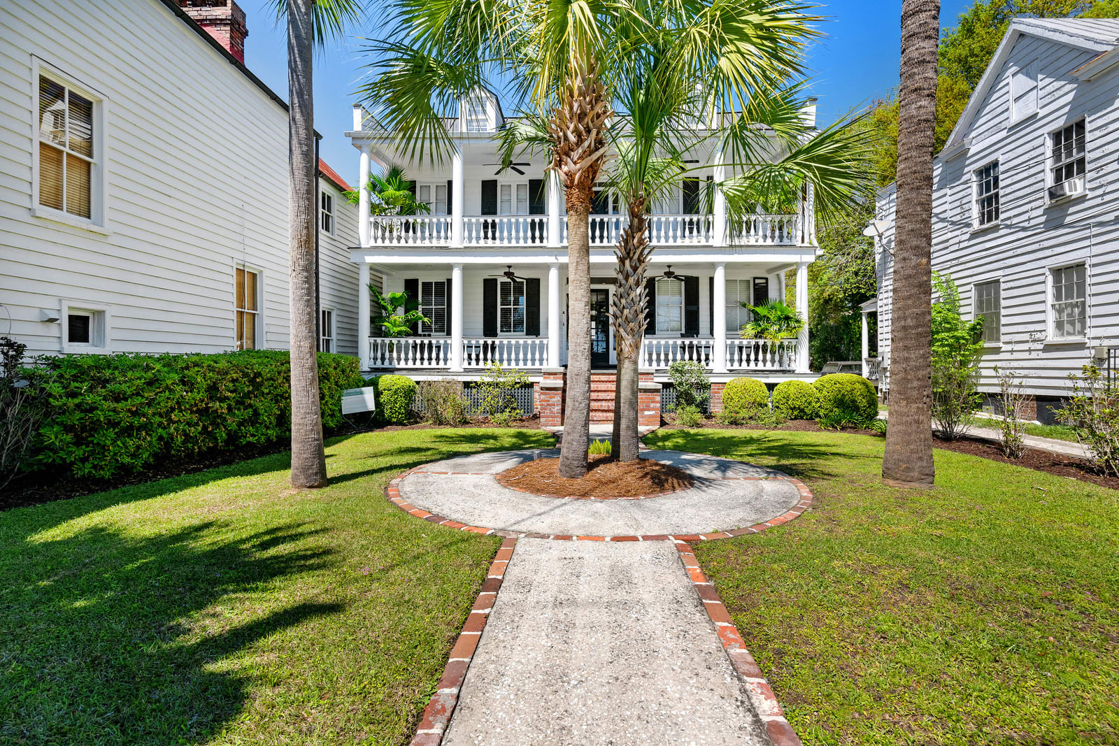 Sunny Southern colonial-style two-story white house with double wraparound porches and columns, palm trees centered in a circular brick-edged walkway, manicured green lawn and neighboring homes