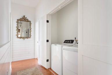 Bright coastal-style hallway laundry nook with white top-load washer and matching dryer behind open pocket doors, white shiplap walls, ornate gold-framed mirror, warm hardwood floors and a woven rug.
