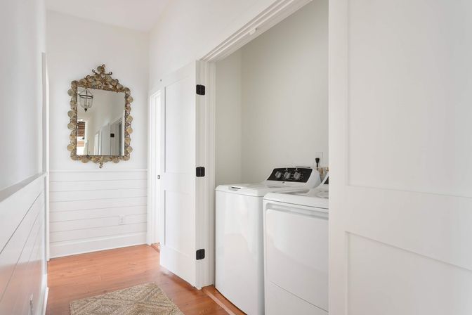Bright coastal-style hallway laundry nook with white top-load washer and matching dryer behind open pocket doors, white shiplap walls, ornate gold-framed mirror, warm hardwood floors and a woven rug.