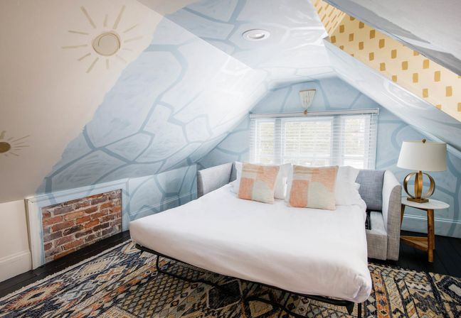 Cozy attic bedroom with sloped ceiling painted pale-blue geometric mural, white pull-out bed with pastel throw pillows, patterned area rug, exposed brick vent, wooden side table and lamp beneath a bright window.