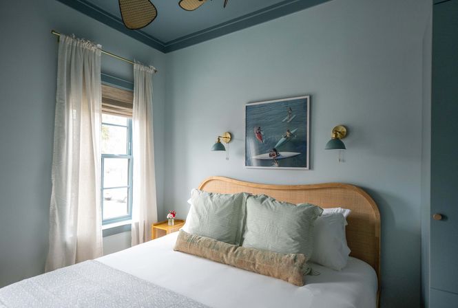 Cozy coastal bedroom with light-blue walls and ceiling, rattan headboard, white bed topped with striped green pillows and a long lumbar cushion, sheer curtains over a window, brass wall sconces and surf-themed artwork.