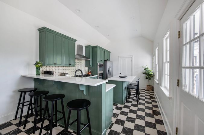 Airy bright kitchen with sage-green cabinets and island, white quartz countertops, stainless steel range and fridge, black bar stools, patterned tile backsplash and black-and-white checkerboard floor under tall windows.