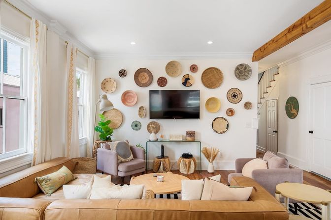 Bright boho-style living room with a tan leather sectional and pastel armchairs around a wooden coffee table, wall-mounted TV framed by an arrangement of woven baskets, large windows and exposed wood beam