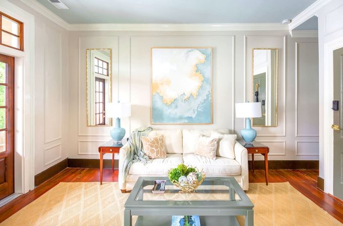 Airy coastal-inspired living room with a cream sofa, patterned throw pillows and a light throw, blue ceramic table lamps on wooden side tables, abstract blue-and-gold wall art, glass-top coffee table with decorative bowl, beige area rug and warm hardwood floors by a sunlit entry door.
