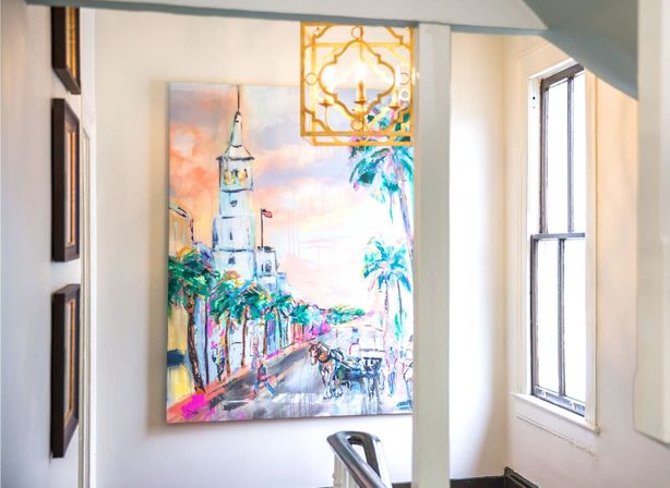 Vibrant coastal street painting of a white tower, palm trees and horse-drawn carriage displayed in a sunlit stairwell beneath a decorative brass pendant light