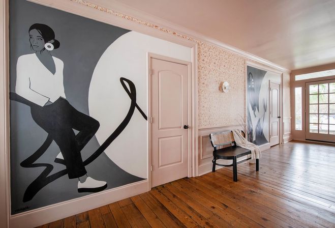 Stylish interior entryway with warm hardwood floors, pastel-pink doors and trim, a large black-and-white mural of a seated woman with flowing ribbon-like lines, a woven bench with a draped throw, patterned wallpaper and a glass-paneled door letting in natural light.