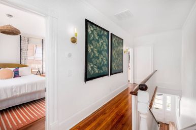 Bright upstairs hallway with polished hardwood floors, framed green botanical prints and brass wall sconces; open doorway reveals a bedroom with white bed, striped rug and rattan pendant light.