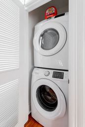 Space-saving stacked white front-load washer and dryer in a compact laundry closet with hardwood floor and a red laundry detergent bottle on top