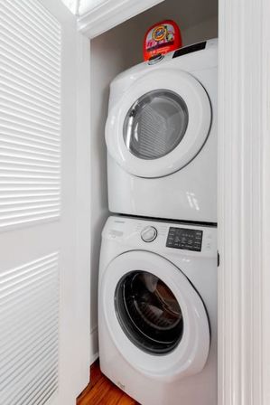 Space-saving stacked white front-load washer and dryer in a compact laundry closet with hardwood floor and a red laundry detergent bottle on top