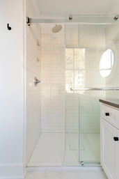 Bright, airy modern bathroom: white subway-tiled walk-in shower with frameless sliding glass door, overhead rain showerhead, recessed niche, and adjacent white shaker vanity with black pulls and natural light from a round window.