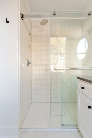 Bright, airy modern bathroom: white subway-tiled walk-in shower with frameless sliding glass door, overhead rain showerhead, recessed niche, and adjacent white shaker vanity with black pulls and natural light from a round window.