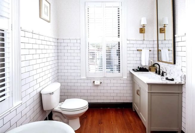 Light-filled white bathroom with subway tile walls, warm wood floor, shuttered window, marble-topped vanity with brass sconces and black faucet, and a toilet.