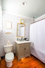 Cozy modern bathroom with white subway tile walls, wood plank floor, gray vanity with marble countertop and black faucet, gold-framed mirror and round brass sconce, white shower curtain and stacked toilet paper on the toilet.