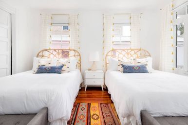 Bright sunlit boho-style twin bedroom with white linens, woven rattan headboards, blue patterned throw pillows, a central white nightstand and lamp, hardwood floors and a colorful geometric rug.