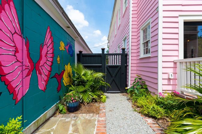 Sunny narrow courtyard with a pink-sided house on the right and a teal mural of pink butterflies and hummingbirds on the left, gravel path to a black gate framed by tropical potted plants.