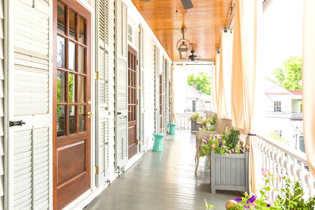 Sunlit covered porch with wooden ceiling, hanging lanterns and fans, glass‑paneled doors with shutters, wicker chairs and colorful potted flowers along a white balustrade.