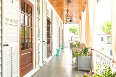 Sunlit covered porch with wooden ceiling, hanging lanterns and fans, glass‑paneled doors with shutters, wicker chairs and colorful potted flowers along a white balustrade.