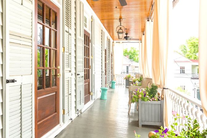 Sunlit covered porch with wooden ceiling, hanging lanterns and fans, glass‑paneled doors with shutters, wicker chairs and colorful potted flowers along a white balustrade.