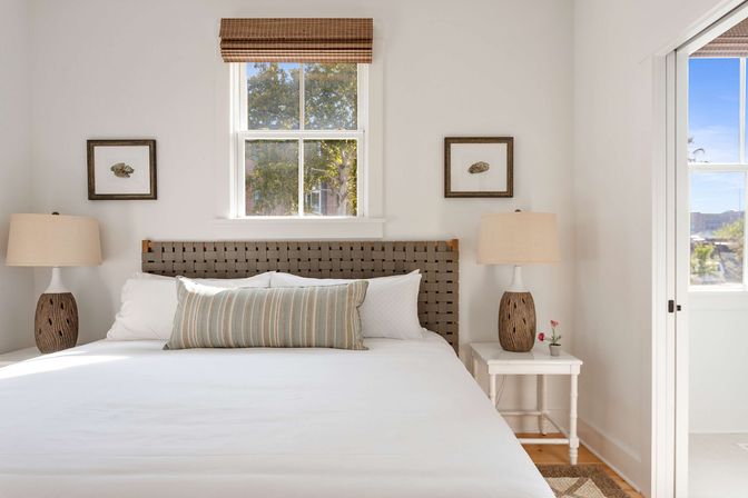 Sunlit coastal-style bedroom interior with white linens, woven headboard, striped lumbar pillow, matching wood-texture bedside lamps and framed shell prints beneath a window