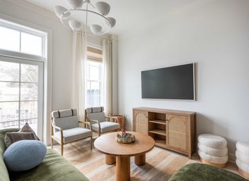 Bright modern living room with large window and wall-mounted TV, natural wood rattan console, round oak coffee table, two light-upholstered chairs, green sofa and textured poufs.