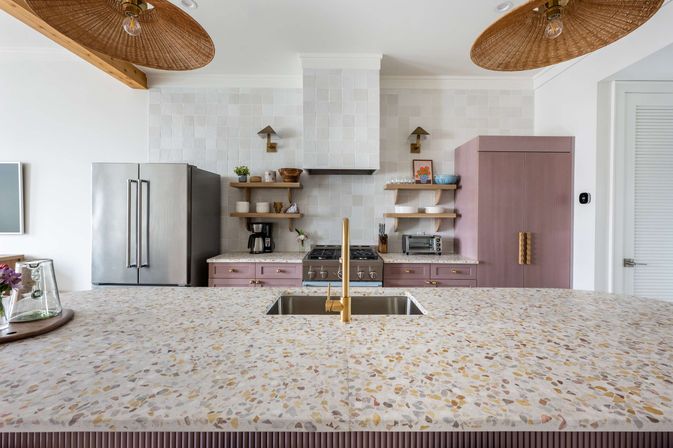 Bright coastal-inspired modern kitchen with large terrazzo island, brass faucet over stainless sink, mauve cabinets, stainless refrigerator, open wood shelves, tiled backsplash and woven pendant lights.