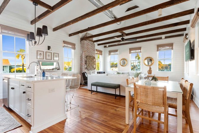 Sunlit coastal open-plan living room and kitchen with exposed dark wood beams, white shiplap walls, hardwood floors, a central island with bar stools, woven dining chairs at a glass table, brick fireplace, and large windows framing palm trees and waterfront views.