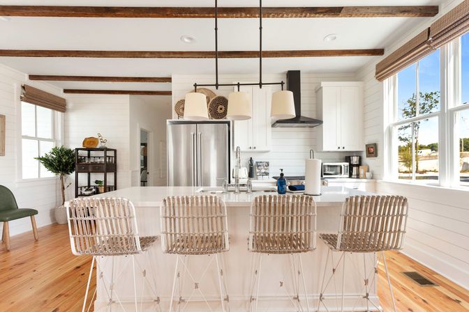 Bright sunlit farmhouse kitchen with white shiplap walls and exposed wood beams, large quartz island with four rattan bar stools, pendant lights, stainless steel refrigerator, black range hood, hardwood floors and big windows overlooking trees.