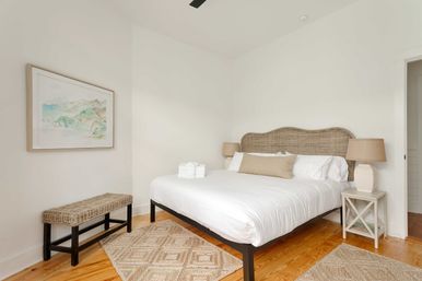 Bright cozy bedroom with a woven rattan headboard and large bed in crisp white linens with a beige lumbar pillow, matching table lamps on white nightstands, a wicker bench, patterned jute rugs on hardwood floors, and soft coastal watercolor wall art.