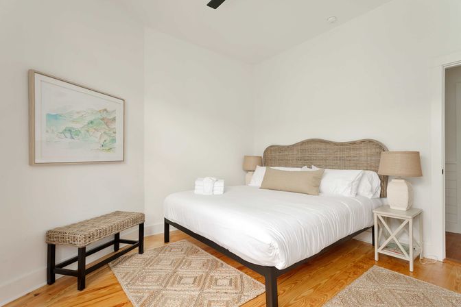 Bright cozy bedroom with a woven rattan headboard and large bed in crisp white linens with a beige lumbar pillow, matching table lamps on white nightstands, a wicker bench, patterned jute rugs on hardwood floors, and soft coastal watercolor wall art.