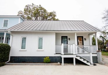 White single-story bungalow with grey metal roof, covered front porch and railing, small shrub and concrete driveway in a quiet residential neighborhood