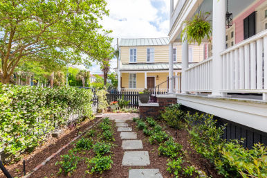 Sunny front garden with stone stepping-stone path, low shrubs and jasmine hedge leading past a white-railed porch with hanging fern to a pastel yellow two-story house and picket fence.