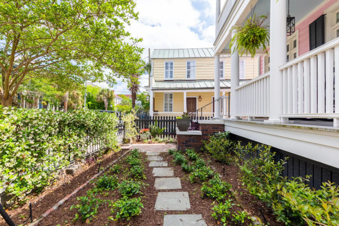 Sunny front garden with stone stepping-stone path, low shrubs and jasmine hedge leading past a white-railed porch with hanging fern to a pastel yellow two-story house and picket fence.