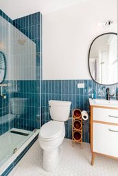 Modern bathroom with vertical teal subway tiles, glass shower, white toilet, hexagon floor tiles, wood-framed vanity and round mirror.