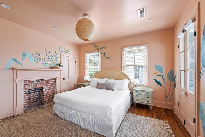 Sunlit peach bedroom with white queen bed, rattan pendant light, exposed brick fireplace, blue botanical wall murals, wooden floors and vintage bedside tables.