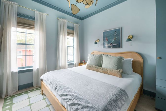 Sunlit coastal bedroom with pale blue walls and ceiling, rattan bed dressed in white linens and green striped pillows, sheer curtains over twin windows, surf art and brass wall sconces.