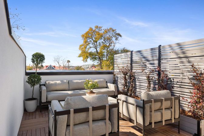 Sunlit rooftop terrace with modern gray outdoor sofa and armchairs on a wooden deck, potted topiary and planters with climbing vines by a slatted privacy screen, tree and city rooftops under a blue sky.