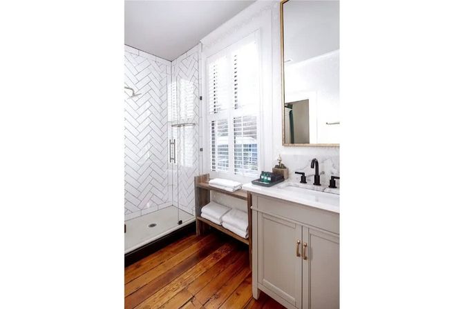 Bright modern bathroom with herringbone white subway-tiled walk-in shower and glass door, warm hardwood floors, marble-topped vanity with matte black faucet, gold-trim mirror, and stacked white towels by a window with plantation shutters.