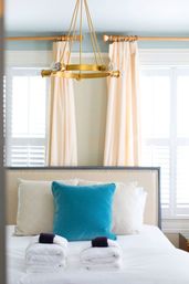 Cozy modern guest bedroom with a brass circular chandelier above a neatly made bed, teal accent pillow, cream headboard, rolled white towels, peach curtains and white plantation shutters.