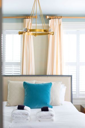 Cozy modern guest bedroom with a brass circular chandelier above a neatly made bed, teal accent pillow, cream headboard, rolled white towels, peach curtains and white plantation shutters.