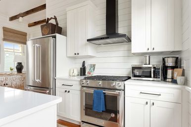 Bright modern farmhouse kitchen with white shiplap walls and exposed wood beams, stainless steel French-door refrigerator and gas range with black vent hood, microwave, coffee maker and toaster on white quartz counters — coastal cottage style.