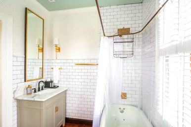 Bright white subway-tile bathroom with marble-top vanity and black faucet, gold fixtures and mirror, shower-curtain tub and sunlit shuttered window.
