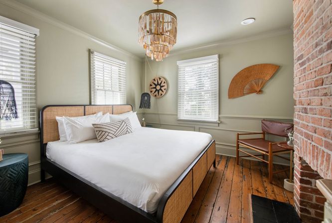 Sunlit neutral bedroom with rattan-framed bed and white linens, hardwood plank floors, exposed brick wall, woven wall fan, pendant chandelier, and leather accent chair.