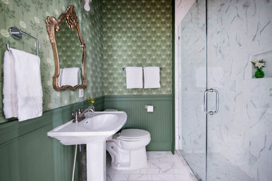 Vintage-style bathroom with green floral wallpaper and beadboard, white pedestal sink and toilet, ornate gold mirror, white towels, and marble-tiled glass shower
