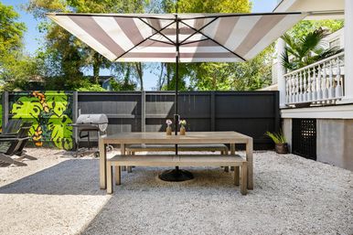 Sunny backyard patio with a wooden dining table and benches under a large striped umbrella on gravel, black fence with tropical leaf mural, charcoal grill and potted plants for al fresco dining.