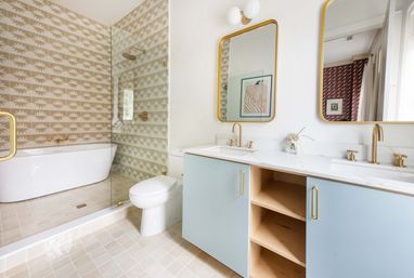 Light-filled modern bathroom with powder-blue double-sink vanity, marble countertop and gold fixtures, glass-enclosed shower, freestanding white tub and beige sunburst-patterned wall tiles