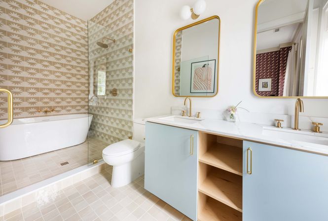 Light-filled modern bathroom with powder-blue double-sink vanity, marble countertop and gold fixtures, glass-enclosed shower, freestanding white tub and beige sunburst-patterned wall tiles
