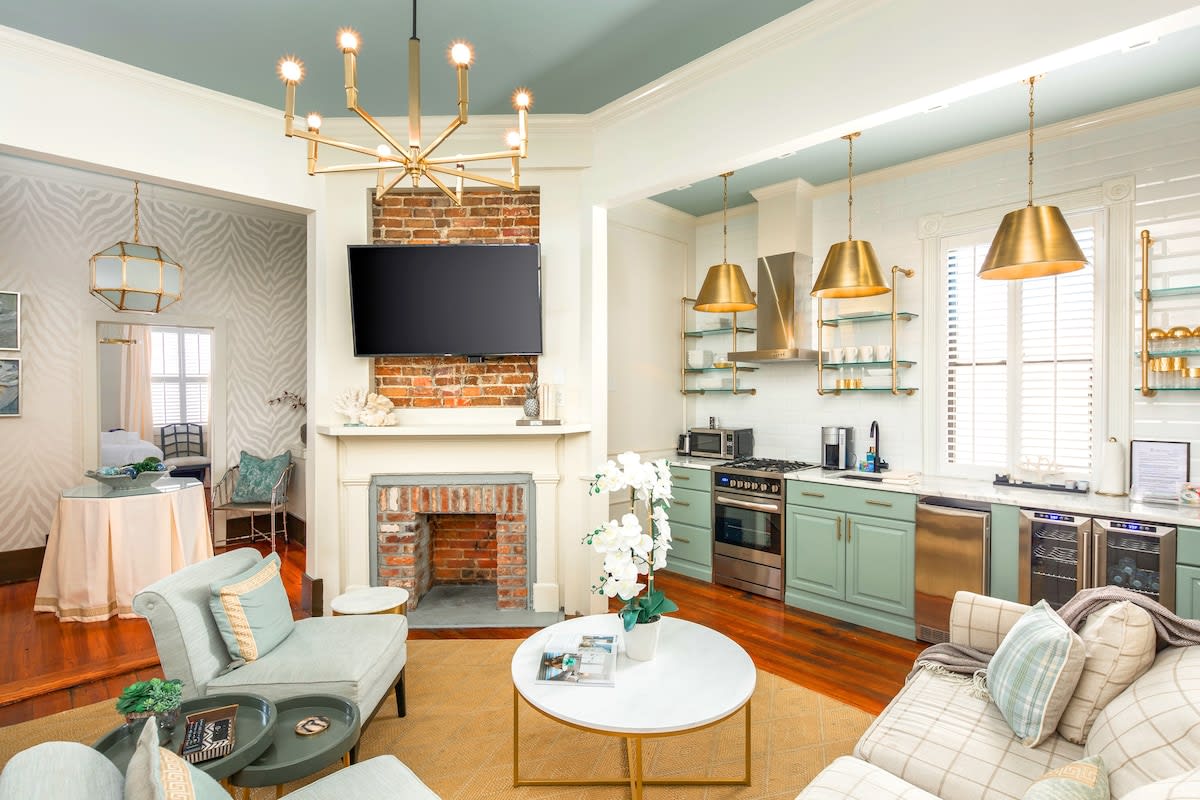 Bright open-concept living room and kitchen with mint-green cabinets, brass pendant lights, exposed brick fireplace with wall TV, light-blue ceiling, hardwood floors, round white coffee table with orchids, pale plaid sofa and cozy seating.