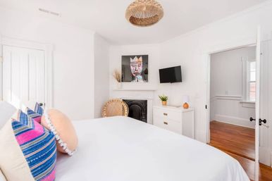 Bright white bedroom with a bed dressed in colorful striped and round pillows, rattan pendant light, decorative black fireplace, wicker chair, wall-mounted TV, white dresser and hardwood floors.