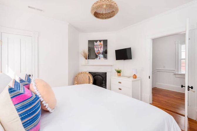 Bright white bedroom with a bed dressed in colorful striped and round pillows, rattan pendant light, decorative black fireplace, wicker chair, wall-mounted TV, white dresser and hardwood floors.