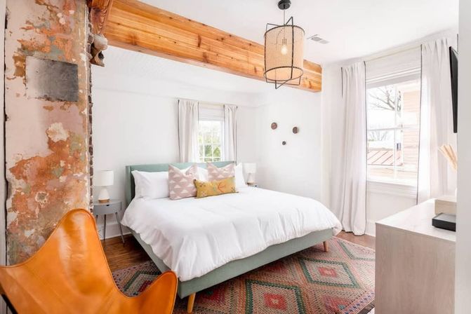 Sunlit modern farmhouse bedroom in an urban loft with exposed wood beam and distressed plaster column, king bed with white duvet, pastel throw pillows, patterned rug, orange leather accent chair and woven pendant light
